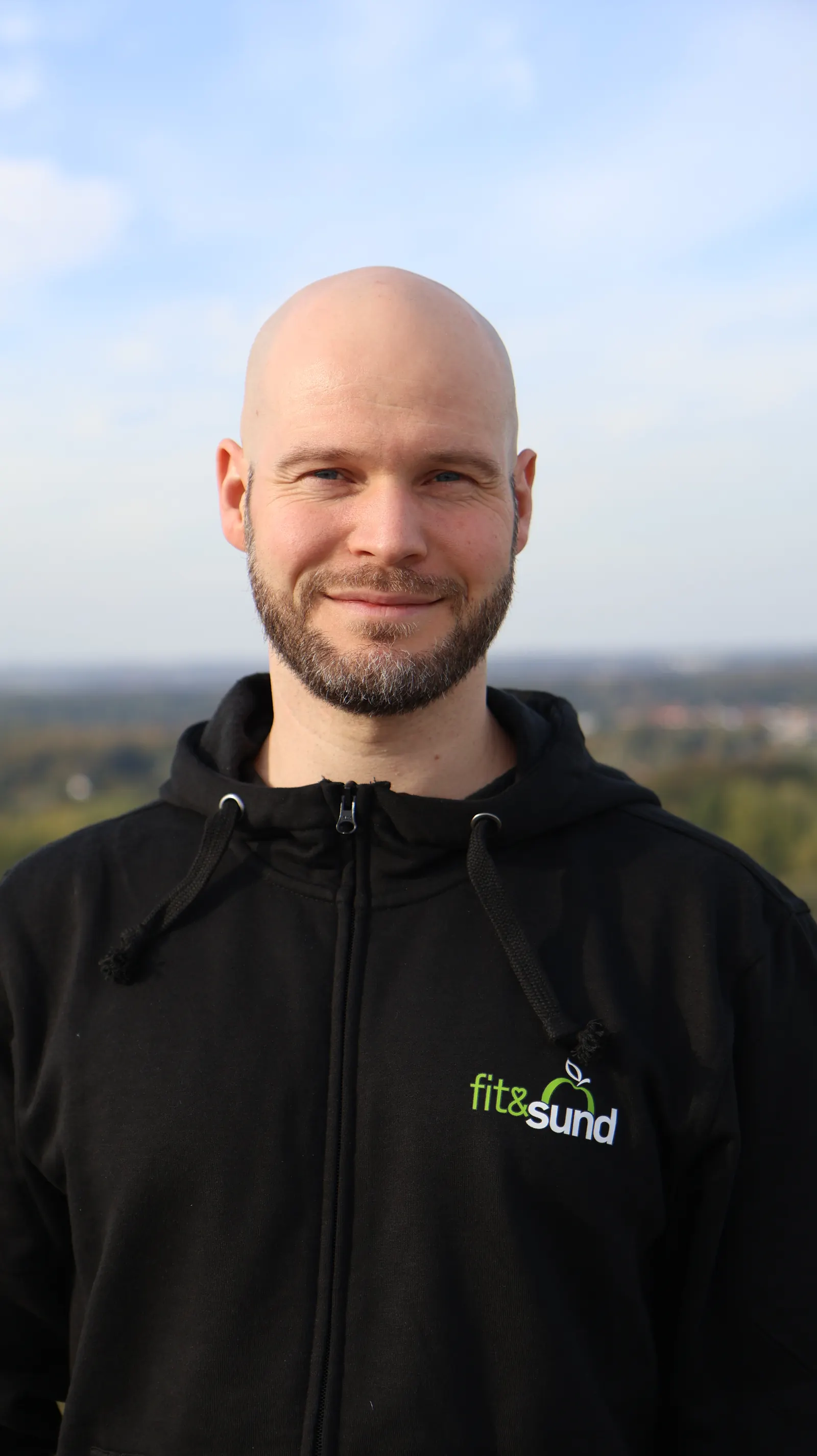 Portræt af fysioterapeut David Brink, der smiler udendørs med himmel og landskab bag sig.