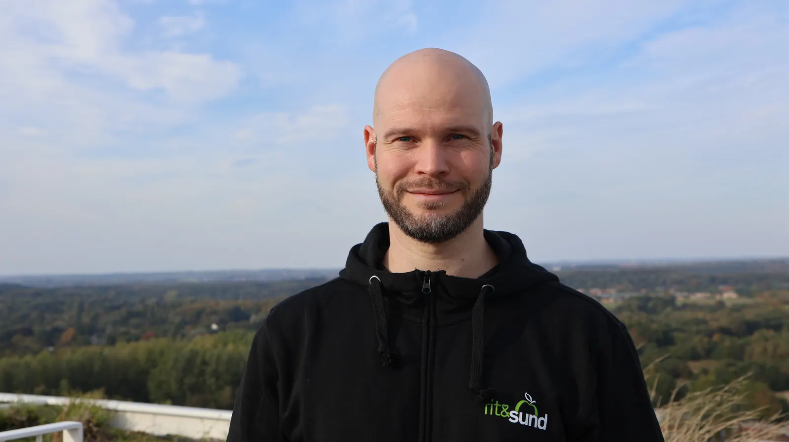 David Brink smiler udendørs med udsigt over skov og by.